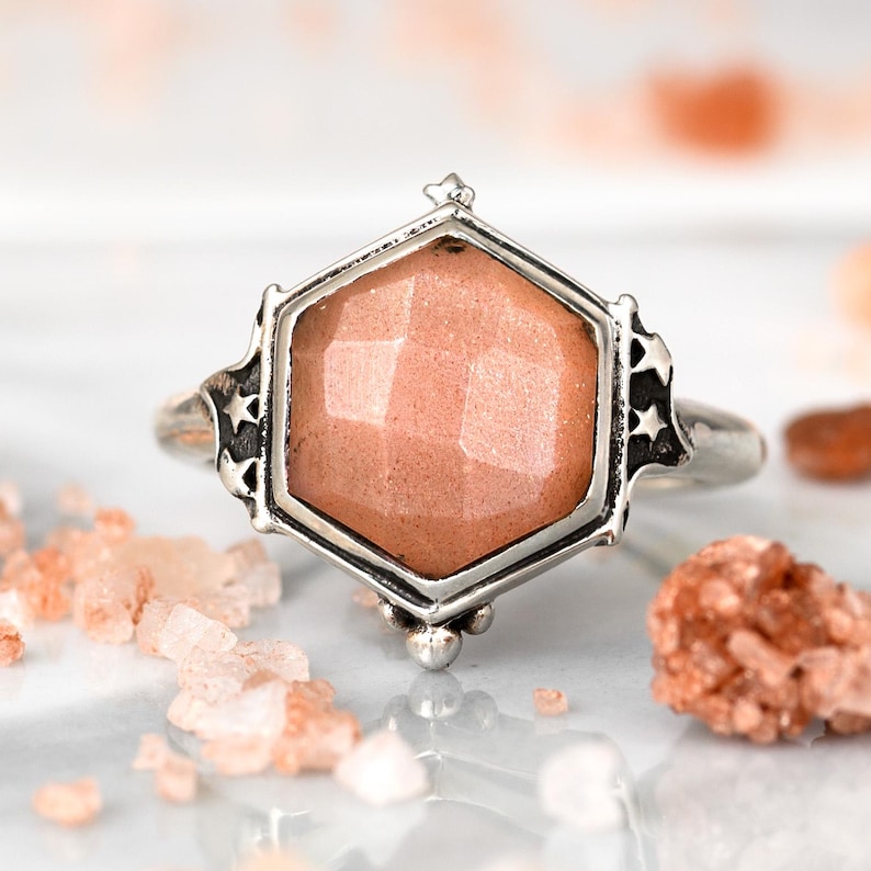 Op de afbeelding: Een zilveren ring met een zeshoekige zetting met een perzikkleurige edelsteen. De ring heeft een stervormig ontwerp op de band.