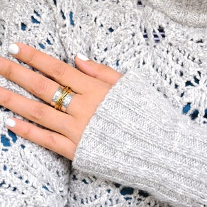 May include: Close-up of a hand with white nail polish wearing multiple rings. The rings are silver and gold, with a textured design. The hand is resting on a gray, open-knit sweater with a blue underlayer.