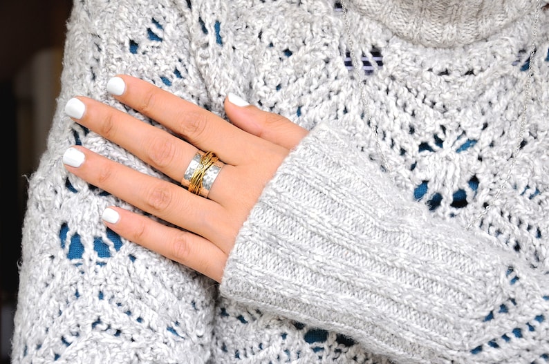 May include: Close-up of a hand with white nail polish wearing multiple rings. The rings are silver and gold, with a textured design. The hand is resting on a gray, open-knit sweater with a blue underlayer.