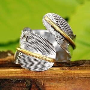 May include: A silver ring with a gold feather design. The feather is wrapped around the ring band and has a textured surface.