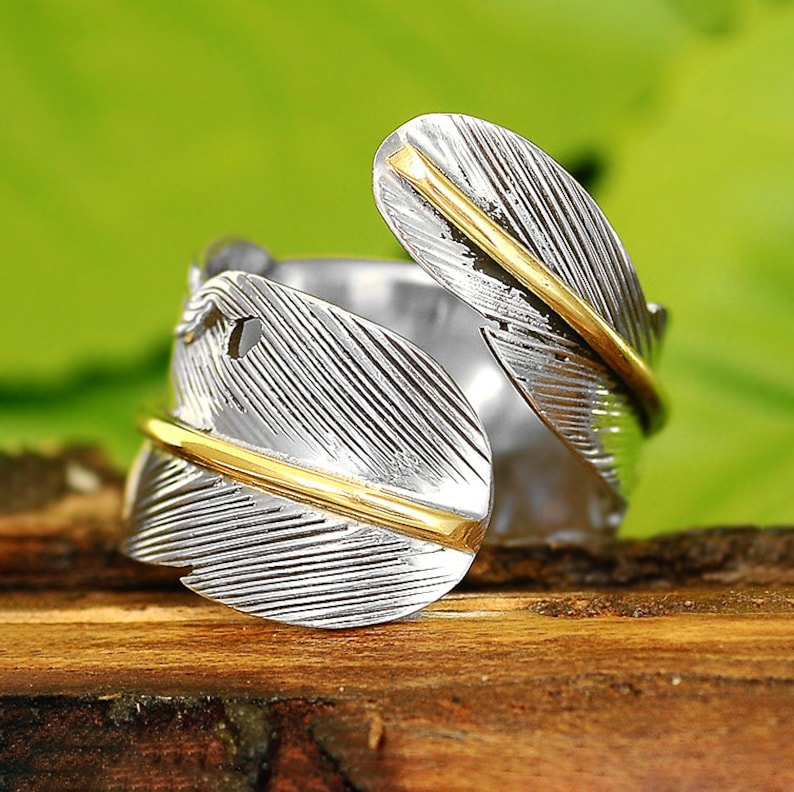 May include: A silver ring with a gold feather design. The feather is wrapped around the ring band and has a textured surface.