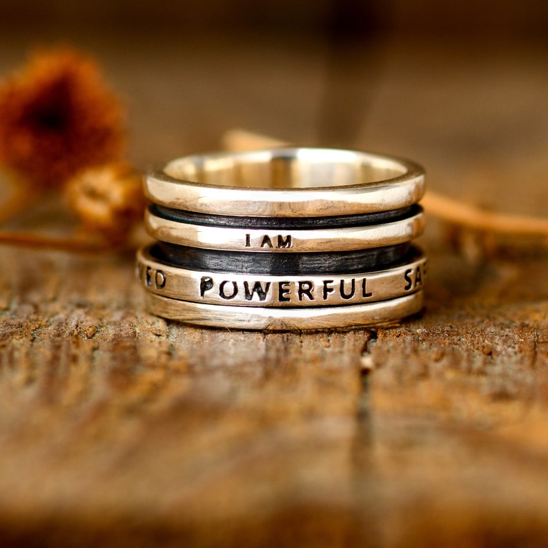 May include: A silver ring with four bands, each engraved with a different word. The words are "I AM", "POWERFUL", "SAFE", and "LOVED".