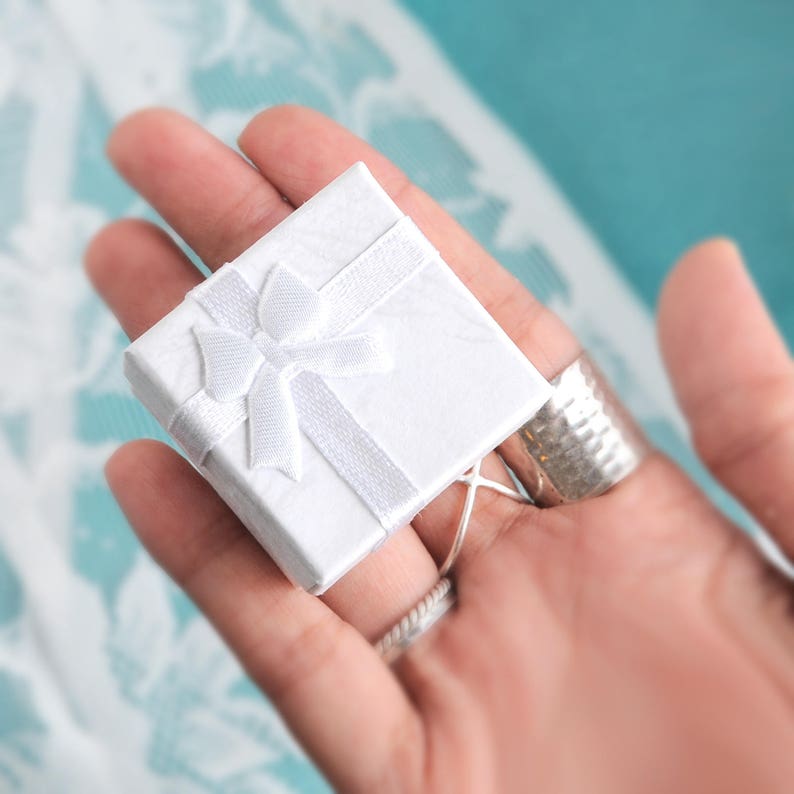 May include: A small, square white gift box with a matching ribbon and bow. The box is held in a hand, with silver rings on the fingers. The background is a soft, light blue.