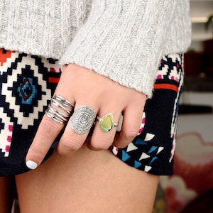 May include: A hand wearing a silver ring with a circular design and a silver ring with a teardrop-shaped green stone.