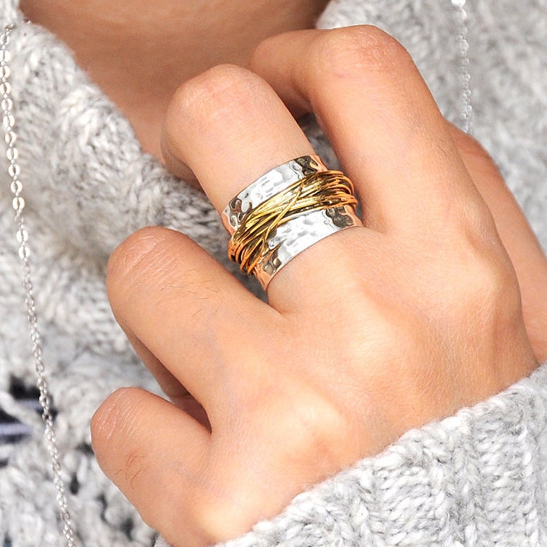 May include: A silver ring with a hammered texture and a gold wire wrap design. The ring is on a finger, and the person is wearing a gray sweater. The ring is a piece of jewelry.