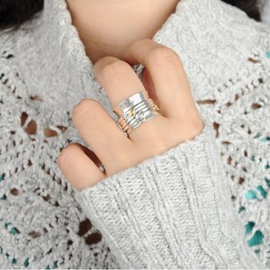 May include: A silver spinning ring with a clear stone and gold accents. The ring is worn on a hand with a grey sweater.