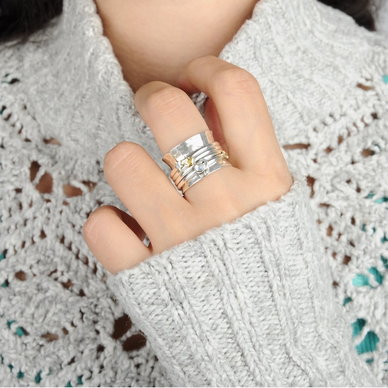 May include: A silver spinning ring with a clear stone and gold accents. The ring is worn on a hand with a grey sweater.