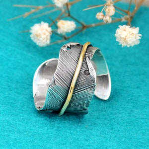 May include: A silver ring with a feather design and a gold band. The feather is textured and has a black stone accent.