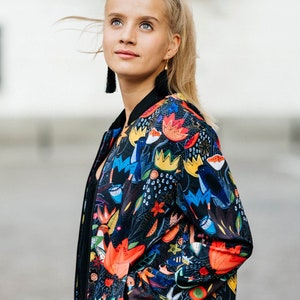 Könnte beinhalten: Schwarze Bomberjacke mit einem lebendigen Blumenmuster in Rot, Orange, Gelb und Blau. Die Jacke hat einen schwarzen Kragen, Bündchen und Saum und verfügt über zwei Taschen. Das farbenfrohe Design bildet einen auffälligen Kontrast zum schwarzen Hintergrund.