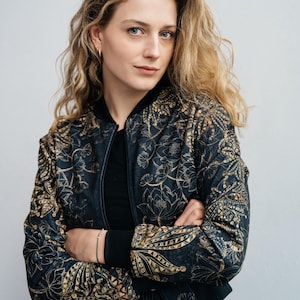 May include: A woman wearing a black bomber jacket with a gold floral pattern. The jacket has a black collar and cuffs. The woman has long, wavy blonde hair and is looking at the camera.