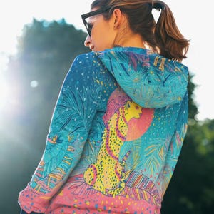 Può includere: Una felpa con cappuccio dal design vivace e colorato. La felpa presenta un'illustrazione di ghepardo con un sole rosa e fogliame tropicale nelle tonalità di blu, rosa, giallo e verde. Anche le maniche e il cappuccio sono decorati con un motivo simile.