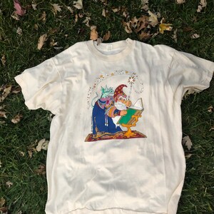 May include: A white t-shirt with a colorful illustration of a wizard with a book and a cat. The wizard is wearing a blue robe and a red hat. The cat is wearing a purple robe and a green hat. The illustration is surrounded by stars.