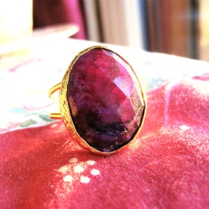 Peut inclure: Une bague dorée ornée d'un grand rubis ovale facetté. Le rubis présente des nuances de rouge profond et de violet. Le bandeau de la bague a un motif texturé. La bague est posée sur un tissu rouge. Le rubis mesure environ 2,5 cm de long.