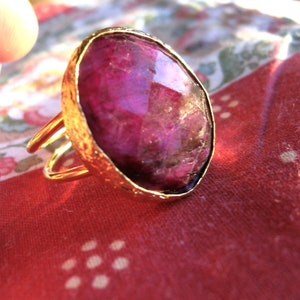 Peut inclure: Une bague dorée avec une grosse pierre précieuse rose foncé, de forme ovale et facettée. La bague a un anneau doré texturé et un design à double bande. La bague est posée sur un tissu rouge à motifs.