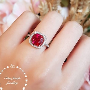 以下が含まれることがあります： 大きな正方形の赤い宝石が、小さな白い宝石のハローで囲まれた銀の指輪です。