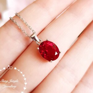 Peut inclure: Un collier en argent avec un pendentif en pierre précieuse rouge vif de forme ovale. La pierre précieuse est sertie dans un sertissage à quatre griffes et est suspendue à une fine chaîne en argent. Le bijou est présenté sur un fond flou.