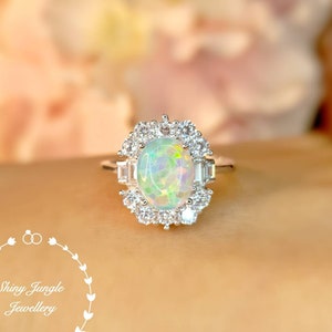 Könnte beinhalten: Ein silberner Ring mit einem großen ovalen Opal-Edelstein, der von kleineren klaren Edelsteinen umgeben ist. Der Ring ist in einem zarten, floral inspirierten Design gehalten.