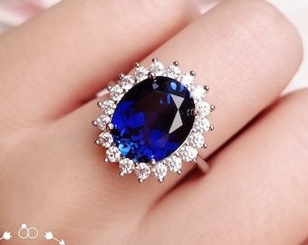 Könnte beinhalten: Ein silberfarbener Ring mit einem großen, oval geschliffenen, tiefblauen Mittelstein, der von einem Kranz aus kleinen, klaren, rund geschliffenen Steinen umgeben ist. Der Text "Shiny Jungle Jewellery" ist unten links mit einer floralen Umrandung zu sehen.