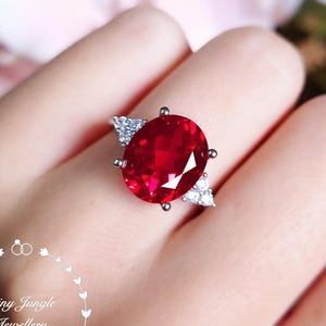 May include: A silver-toned ring featuring a large, oval-cut vibrant red gemstone at the center, flanked by two small triangular clusters of clear white stones on each side. The text Shiny Jungle Jewellery is visible in a circular frame.