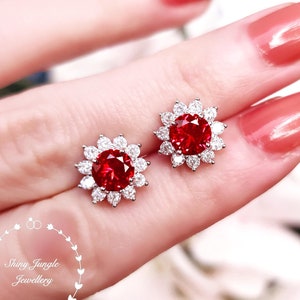 May include: A pair of stud earrings featuring a large, round, red gemstone surrounded by smaller, clear gemstones in a floral design. The earrings are set in a silver-colored metal. The image also includes the text "Shiny Jungle Jewellery".