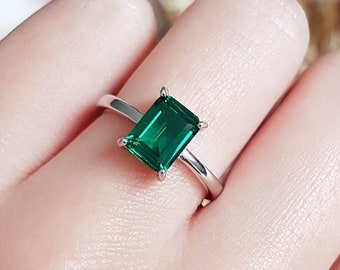 May include: A silver-toned ring featuring a rectangular emerald-cut green gemstone held by four prongs. The band is smooth and polished. A circular logo with the text Shiny Jungle Jewellery is visible in the lower right corner of the image.