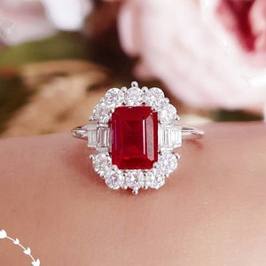 May include: A silver ring featuring a large, emerald-cut red gemstone encircled by a halo of smaller, round white gemstones.