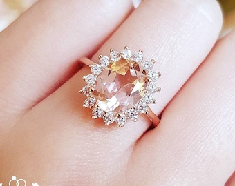 May include: A rose gold ring featuring a central oval-cut peach-colored gemstone surrounded by a halo of small round white stones. The band is thin and metallic. The text Shiny Jungle Jewellery is visible in the lower left corner.