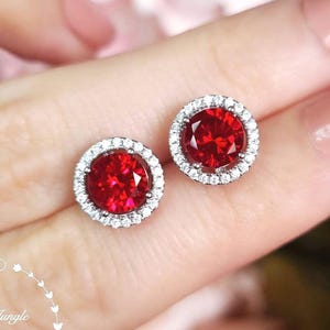 Puede incluir: Un par de pendientes de botón redondos con una piedra central de color rojo vibrante rodeada por un halo de pequeñas piedras blancas transparentes. El texto Shiny Jungle Jewellery aparece en la esquina inferior izquierda.