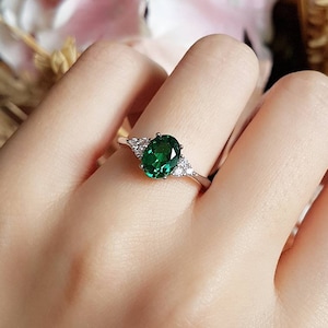 May include: A silver ring featuring an oval-cut, vibrant green gemstone as the centerpiece. Two smaller, clear gemstones flank the main stone. The ring is set on a delicate band, showcasing a classic and elegant design.