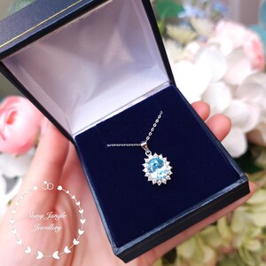 May include: A silver necklace with a large, oval, light blue gemstone pendant surrounded by smaller white gemstones, presented in a dark blue jewelry box. The box is open, revealing the necklace against a velvet interior. The text "Shiny Jungle Jewellery" is visible.