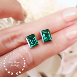 May include: A pair of emerald-cut, green gemstone stud earrings. The earrings are set in a silver-colored metal. The image includes the text "Shiny Jungle Jewellery" in a circular design.