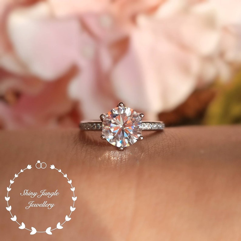 May include: A silver-toned ring featuring a large, round, faceted center stone held by six prongs, with smaller stones embedded along the band. The text Shiny Jungle Jewellery is visible in the lower left corner within a floral wreath.