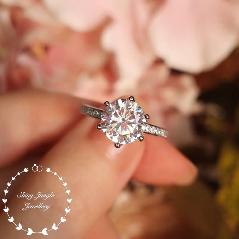 May include: A silver-toned ring featuring a large, round, faceted center stone held by six prongs, with smaller stones embedded along the band. A circular logo with the text Shiny Jungle Jewellery is visible in the lower left corner.