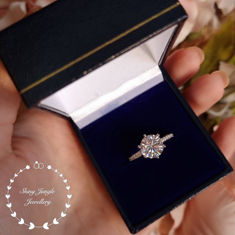 May include: A silver-toned ring featuring a large, round, faceted center stone held by six prongs, with smaller stones set along the band. The ring is presented in a dark blue, hinged jewelry box with a white interior lining and the text Shiny Jungle Jewellery.