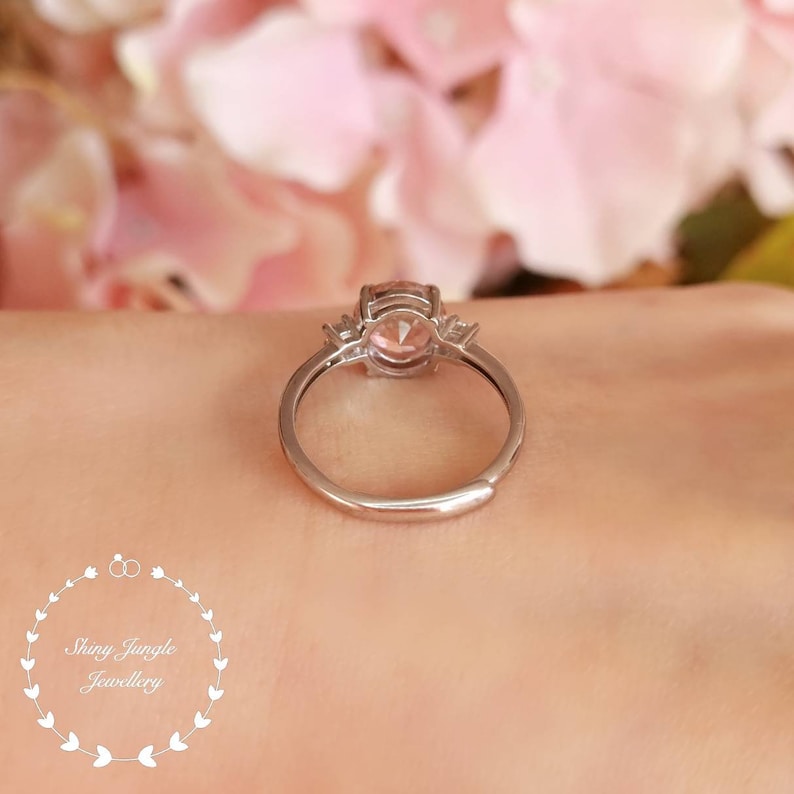May include: A silver ring with a pink gemstone and two smaller clear gemstones on the band.