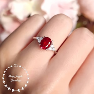 May include: A silver ring featuring a large oval ruby gemstone, surrounded by smaller white gemstones.