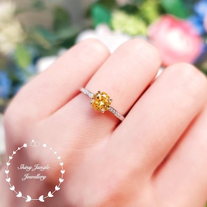 May include: A silver ring featuring a round, yellow gemstone encircled by smaller, clear gemstones. The ring is worn on a finger.