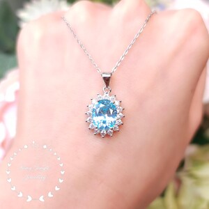 May include: A silver necklace with a large, oval, light blue gemstone pendant. The pendant is surrounded by a halo of smaller, clear gemstones. The necklace is displayed against a soft, blurred background.