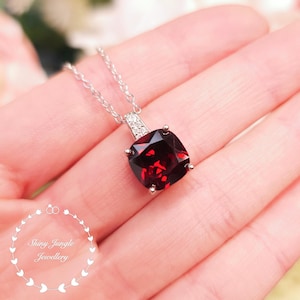 May include: A silver necklace with a square, deep red gemstone pendant. The pendant is set in a silver frame and has a small, diamond-studded bail. The necklace is held in a person's hand. The text "Shiny Jungle Jewellery" is in the lower left corner.