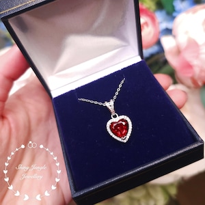 Puede incluir: Un collar de plata con un colgante en forma de corazón adornado con una piedra roja rodeada de piedras transparentes más pequeñas. El collar se muestra en una caja de joyería de terciopelo azul.