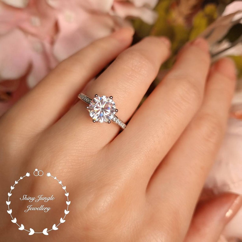 May include: A silver-toned ring featuring a large, round, brilliant-cut clear center stone held by six prongs, with smaller clear stones embedded along the band. The image includes a logo with the text Shiny Jungle Jewellery.