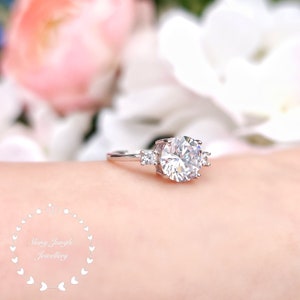 May include: A silver ring with a large, round, clear gemstone surrounded by three smaller, clear gemstones. The ring is on a person's finger.