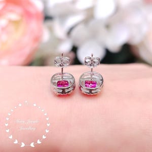 May include: A pair of silver stud earrings with a bright pink gemstone center, surrounded by a halo of smaller stones. The earrings are set against a soft pink background, with a blurred floral backdrop.