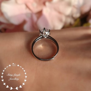 May include: A silver-toned ring featuring a single clear, round-cut stone held in a prong setting. The band is open at the bottom, indicating an adjustable size. The logo for Shiny Jungle Jewellery is visible in the bottom left corner.