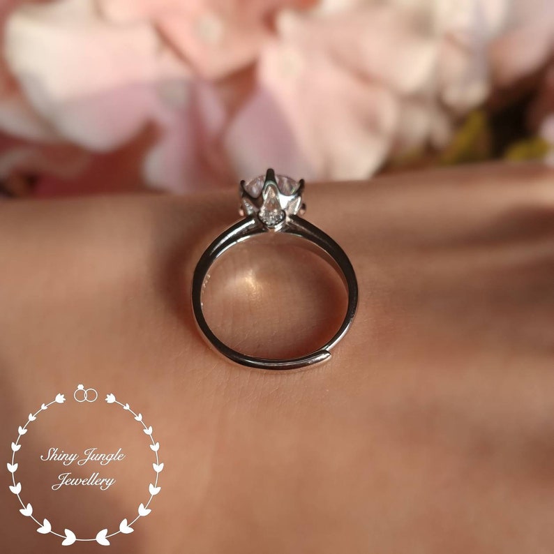 May include: A silver-toned ring featuring a single clear, round-cut stone held in a prong setting. The band is open at the bottom, indicating an adjustable size. The logo for Shiny Jungle Jewellery is visible in the bottom left corner.