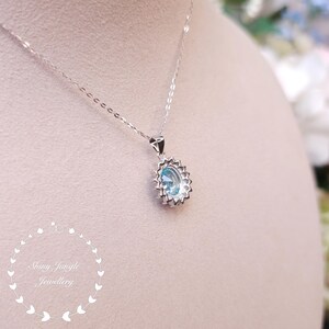 May include: A silver necklace with a pendant featuring a light blue oval gemstone surrounded by small, clear stones. The pendant hangs from a delicate chain. The image also includes the text "Shiny Jungle Jewellery".