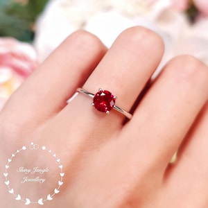 May include: A silver ring featuring a prominent, round, deep red gemstone. The ring is set on a delicate band and is shown on a finger. The image is well-lit, emphasizing the gemstone's colour and clarity. The ring is a piece of jewellery.