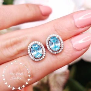 May include: A pair of oval-cut light blue stud earrings, each featuring a central blue stone surrounded by a halo of small clear white stones set in a silver-toned metal. The text Shiny Jungle Jewellery is visible in the lower left corner.