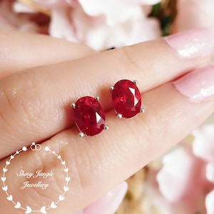 May include: A pair of oval-shaped red gemstone earrings set in silver. The earrings are shown on a person's earlobe.