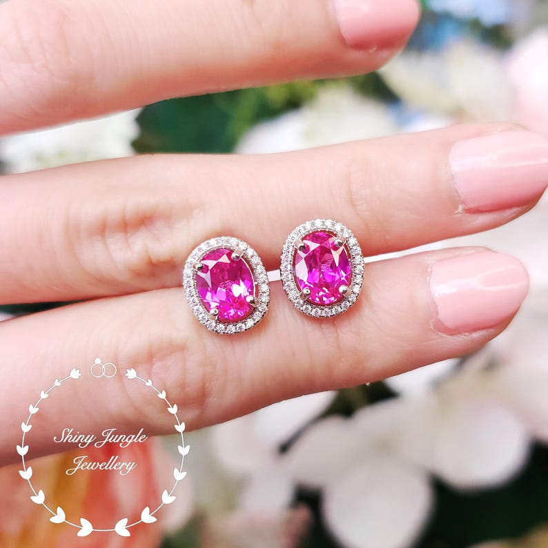 May include: A pair of oval-shaped, pink gemstone stud earrings. Each earring is surrounded by a halo of small, clear gemstones set in a silver-colored metal. The earrings are displayed against a soft, blurred background.